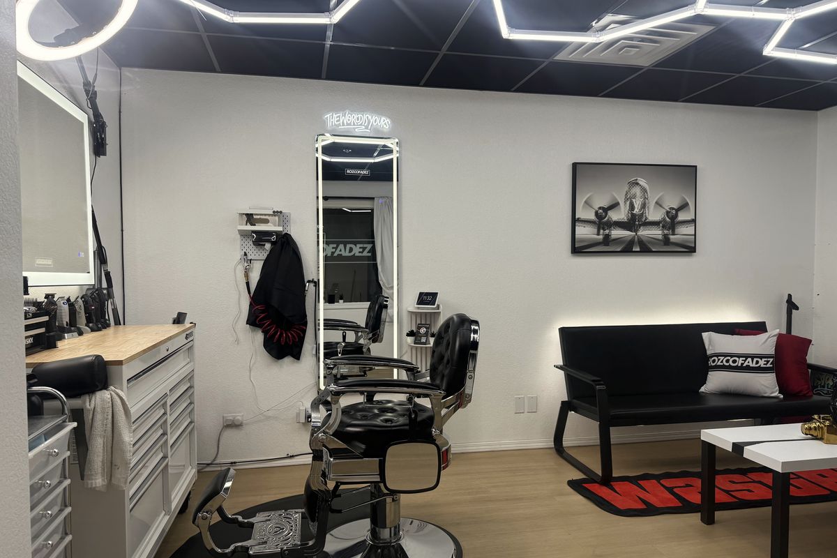 The full Rozco Fadez private studio in Suite 180 at The Barber Hall, Boise: barber chair and tool station on the left, a tall lit mirror under a neon sign, and a black lounge couch, all under hexagon ceiling lights